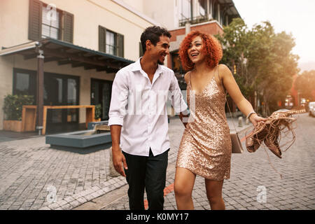 Junge Mann und die Frau zu Fuß auf einer verlassenen Straße. Lächelnde Frau in glamourösen kleiden Sie Ihre Sandalen Halten in der Hand und gehen mit Ihrem Partner. Stockfoto