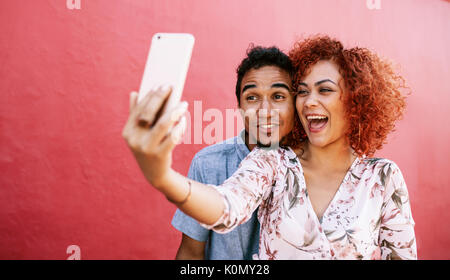 Junge Frau mit einem Smartphone für die selfie mit Ihrem Partner. Glückliches junges Paar über ein Telefon Ihr Foto gegen den roten Hintergrund zu nehmen. Stockfoto