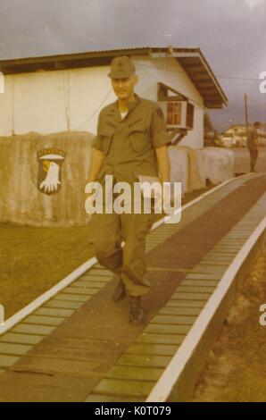 Ein Soldat, als Robert Swisher der 101St Airborne Division, gesehen können Sie zu Fuß zu einem Holzsteg, von aussen ein Gebäude mit einem klimagerät gesehen werden kann, mit einem anderen Soldaten im Hintergrund sein, Camp Eagle, Vietnam, 1964. Stockfoto