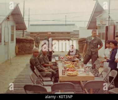 Tet Holiday, amerikanische Soldaten genießen Sie eine Mahlzeit an einem langen Tisch auf den Urlaub an ihrer Basis in Vietnam Feiern während des Vietnam Krieges, Getränke und andere Lebensmittel sichtbar auf dem Tisch, mehrere Südvietnamesischen Frau, die in der Nähe stehen, 1965. Stockfoto