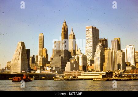 Blick nach Westen von Morgensonne auf Wolkenkratzer in Manhattan, New York City, August, 1966 glänzen. Ein Brand in den East River spritzt Wasser in Richtung der alten Station Linie Docks am Pier 15, 16 und 17, entlang der South Street Seaport Shoreline. Mehrere Financial District Wahrzeichen sind im Hintergrund sichtbar, von Links nach Rechts: Die zikkurat inspirierte 120 Wall Street; der American International Building bei 70 Pine Street; und die Manhattan Company Building 40 Wall Street. Stockfoto