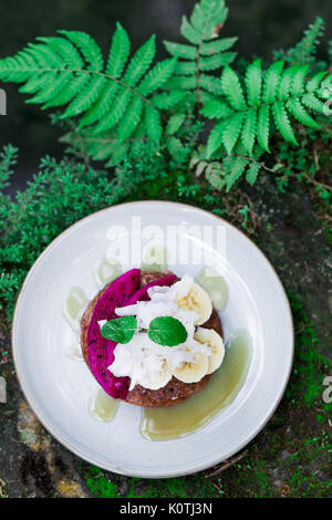 Leckeres Frühstück draußen im Garten. Frische Honig vollkorn Pfannkuchen mit in Scheiben geschnittenen dragon Obst, Bananen, Kokosnuss und Minze. Tasse schwarzen Bali Coffee Stockfoto