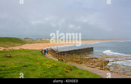 Lange einsame Sandstrand in Seaton Sluice in der Nähe von Dorf an der Küste von Hartley, mit Menschen zu Fuß auf vorland Weg und die Stadt in der Ferne, England Stockfoto