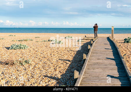 Eine Frau mit Blick aufs Meer mit dem Fernglas vom Ende einer Promenade über dem Strand in Dungeness zu Stockfoto
