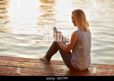 Frauen auf der Suche nach Handy während der Sitzung auf dem Fluss dock Stockfoto