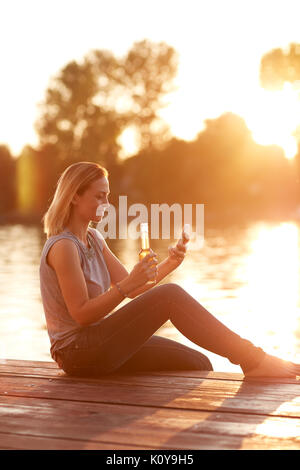 Frau auf der Suche nach Handys auf dem Dock und erfrischt mit Drink bei Sonnenuntergang Stockfoto