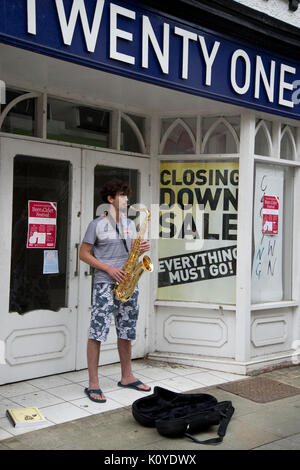 West Wales. Haverford West. Bridge Street. Gaukler spielen ein Saxophon außerhalb einer geschlossen Shop. Stockfoto