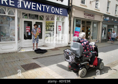 West Wales. Haverford West. Bridge Street. Gaukler spielen ein Saxophon außerhalb einer geschlossen Shop und eine Frau an einem Mobility Scooter. Stockfoto