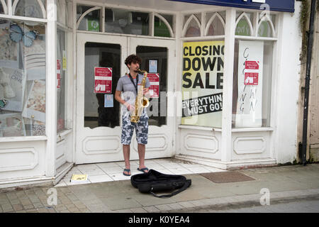 West Wales. Haverford West. Bridge Street. Gaukler spielen ein Saxophon außerhalb einer geschlossen Shop. Stockfoto