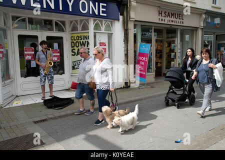 West Wales. Haverford West. Bridge Street. Ein paar Gehminuten hinunter die Straße mit ihren Hunden, während ein strassenmusikant spielt eine Saxophon außerhalb einer geschlossen Shop. Stockfoto