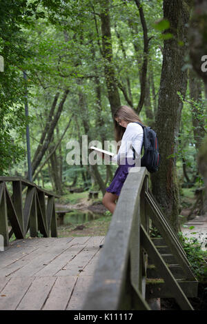 Hübsche kleine blonde Schülerin mit Rucksack stand auf einer Brücke in der Natur und lesen ein Buch fertig, zurück zu Schule studieren für ex zu gehen Stockfoto