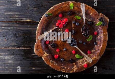 Frische reife Beeren Himbeeren Brombeeren und Heidelbeeren auf braunem Holz- Platte mit einem Löffel auf einer schwarzen Holztisch, rustikal, selektiver Fokus, Spa Stockfoto