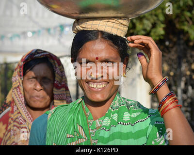 Die indische Adivasi-Frau (Orissan Tribal Woman) mit zwei markanten goldenen Nasenstollen balanciert auf dem Kopf eine schwere Metallschale und lächelt für die Kamera. Stockfoto