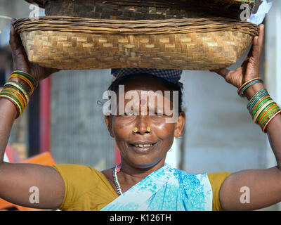 Die ältere indische Adivasi-Frau (Stammfrau) mit zwei markanten goldenen Nasenstollen trägt auf dem Kopf einen Satz Körbe und lächelt für die Kamera. Stockfoto