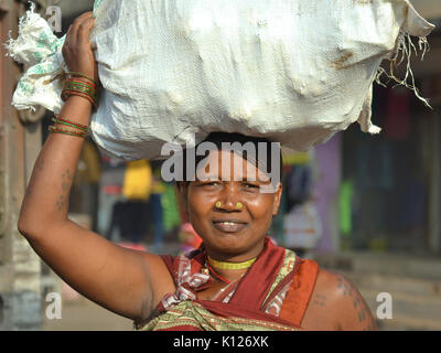 Indische Adivasi-Frau (Stammfrau) mit zwei markanten goldenen Nasenstollen balanciert auf dem Kopf eine schwere Tüte Gemüse und lächelt für die Kamera. Stockfoto