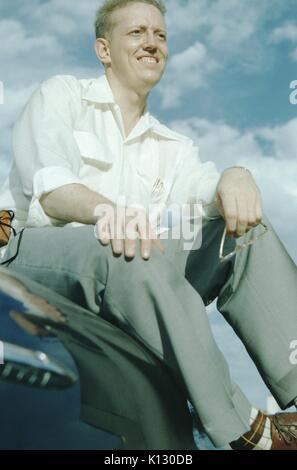 Missionar mit Crew, die auf der Motorhaube eines Autos sitzt, eine Brille hält, Plaid-Socken und Lederschuhe trägt, lächelnd und nach rechts schauend, tiefer Winkel, blauer Himmel mit Wolken im Hintergrund, 1952. Stockfoto