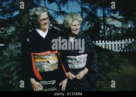 Reifen kaukasischen Frauen, christliche Missionare, tragen Kimonos aus Seide, lächeln und posieren für ein Foto vor der immergrünen Bäumen, beide aus dem Rahmen, Japan, 1951. Stockfoto