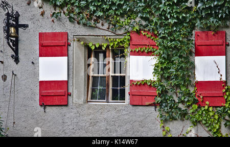 Verputzte Wand mit Fenster, grünen Reben, antike Bell und roten und weißen Fensterläden aus Holz Stockfoto