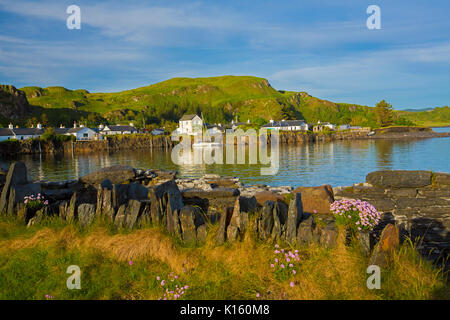 Herrliche Aussicht auf Dorf, Seil Ellenabeich/Easdale Island, Schottland, mit weißen Häuschen an der Basis der felsigen Hügel & neben ruhigen blauen Wasser des Ozeans Stockfoto