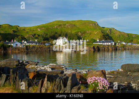 Herrliche Aussicht auf Dorf, Seil Ellenabeich/Easdale Island, Schottland, mit weißen Häuschen an der Basis der felsigen Hügel & neben ruhigen blauen Wasser des Ozeans Stockfoto