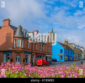 Schottischen Dorf Tarbert, bei der die Zeile der bunten Gebäude/Geschäfte Hauptstraße und lebendige rosa Blumen vorne an der Grenze unter blauem Himmel Stockfoto