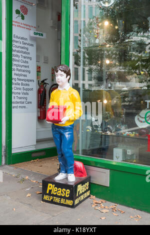 Bitte helfen Sie den Blinden Sammelbox und Statue außerhalb Verhütung von Blindheit der Gesellschaft betrachten wieder Laden auf der Kensington High Street, London, UK Stockfoto