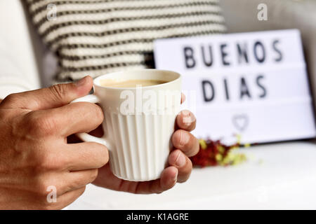 Nahaufnahme von einem jungen Mann mit einem weißen Keramik Tasse mit Kaffee in der Hand und eine Lightbox im Hintergrund mit dem Text Buenos dias, guten Morgen in Sp Stockfoto