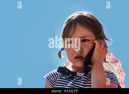 kleines Mädchen mit einem unglücklichen Blick und traurigen Augen Gespräch am Telefon mit meiner Mutter auf dem Hintergrund der schönen blauen Himmel draußen im Park. Stockfoto