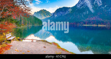 Kontemplative Landschaft mit einer Holzbank auf dem Alpsee Ufer, im Herbst Dekor, mit der Bayerischen Wälder und Berge im See spiegeln Stockfoto