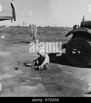 Ein kleiner Junge spielt mit einem handgefertigten Spielzeug Wagen im Schmutz eines Migranten Camp, in der Nähe ein Auto, ein Stück robuste Kleidung trocknet im Hintergrund, Kalifornien, 1936. Von der New York Public Library. Stockfoto