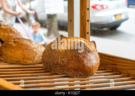 Bronx, NY, USA. Laib des frisch gebackenen gesäte Grieß Brot, angezeigt in einem bakersy Fenster auf einem Holzregal. Street, geparkte Autos und Frau drücken Stockfoto