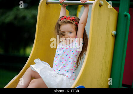 Ein süßes kleines Barfuß girly ist das Spielen auf dem Spielplatz. Baby sitzt auf die Kinder schieben. Auf den Kopf des Mädchens ist ein roter Kranz. Stockfoto