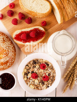 Gesundes Frühstück. Müsli Frühstück mit Müsli, Milch, Marmelade, Brot und frische reife Himbeeren auf einer hölzernen Tabelle, Ansicht von oben Stockfoto