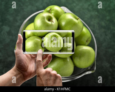 Zwei Hände, die ein mobiles Smartphone und ein Bild von grünen Äpfeln nehmen Stockfoto
