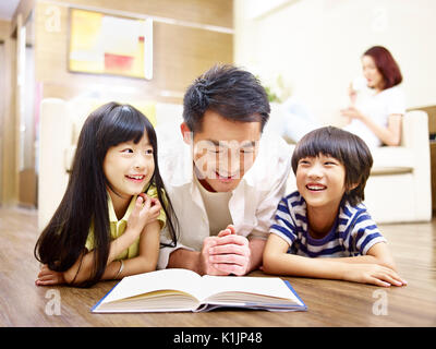 Asiatische Vater und Kinder auf dem Boden lesen Buch liegen mit Mutter im Hintergrund. Stockfoto