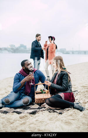 Schöne junge romantisches Paar sitzt auf einem Sandstrand mit Gläsern Wein und einen Korb bei Alfresco Picknick mit unscharfen sprechen Leute auf backgroun Stockfoto