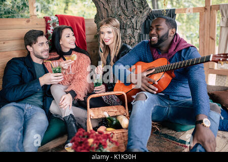 Vier lächelnden jungen Freunde Gläser Holding mit Cocktails und Gitarre an Picknick geniessen. Stockfoto