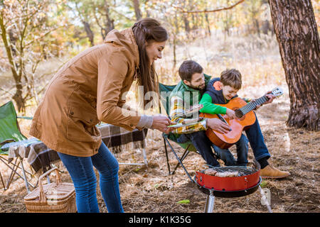 Lächelnd Vater seinen kleinen Sohn, Gitarre zu spielen und die brünette Frau grillen Barbecue Grill im Herbst park Lehre. Happy Family auf Picknick Stockfoto