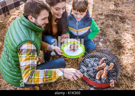 Fröhliche Eltern mit niedlichen kleinen Sohn grillen Würstchen auf Grill im Herbst Park Stockfoto