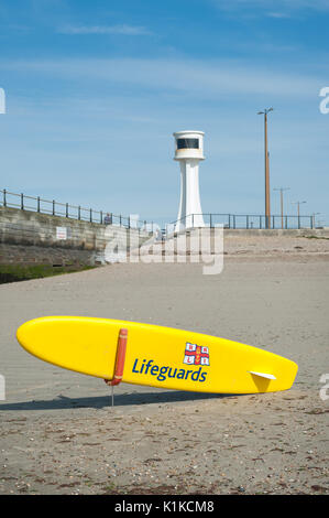 Eine gelbe RNLI Rettungsschwimmer paddle Board oder Surfboard vor einem Leuchtturm auf einem sandigen Strand bei Ebbe in Littlehampton, West Sussex, England. Stockfoto