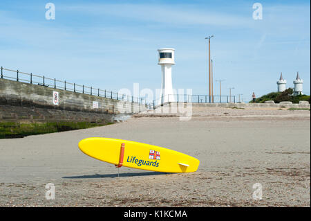 Eine gelbe RNLI Rettungsschwimmer paddle Board oder Surfboard vor einem Leuchtturm auf einem sandigen Strand bei Ebbe in Littlehampton, West Sussex, England. Stockfoto