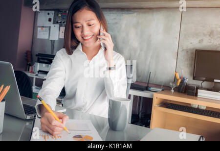 Asiatische Geschäftsfrau mit lächelnden Gesicht sprechen auf mobilen und Schreiben von Daten auf Blatt auf dem Schreibtisch mit Kollegen am Telefon analysieren, Büro Leben anhand von quantitativen Simulatio Stockfoto