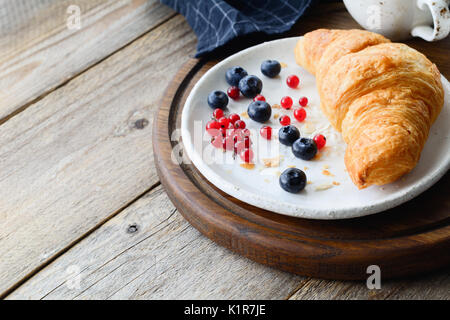 Frische Croissants, Heidelbeeren, rote Johannisbeeren auf weißen Teller und Tasse Kaffee auf Holz- Fach. Kontinentales Frühstück Stockfoto