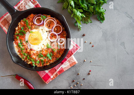 Shakshuka in einer gusseisernen Pfanne. Mitten im orientalischen Stil Eier in tomaten und paprika sauce mit Petersilie, Zwiebeln und Gewürzen auf grauem Beton Hintergrund pochiert. Stockfoto