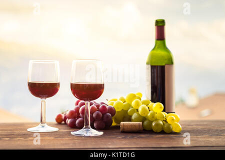 Zwei Gläser guten Rotwein mit Trauben, Flasche und Korken auf einen hölzernen Tisch während einer romantischen Sommer outdoor Diner Stockfoto