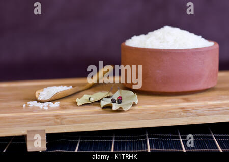 Weißer Reis in eine Schüssel und Lehm in den hölzernen Löffel. Lorbeer und schwarze Pfefferkörner. Stockfoto