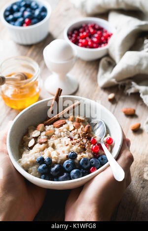 Haferflocken porridge Bowl mit Superfoods in Händen. Hände halten Schüssel Haferflockenbrei mit Blaubeeren, Mandeln, Leinsamen, Zimt und Johannisbeeren. Selectiv Stockfoto