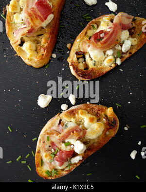 Drei Toast crustinis mit Prosciutto, Kräuter und Käse auf einem schwarzen Holz Oberfläche Stockfoto