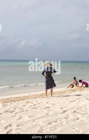 Asiatische Frau mit Schlapphut und einfachen 3/4-Ärmel Kleid auf den Hüften an stürmischen Karibik suchen. Von hinten gesehen. Stockfoto