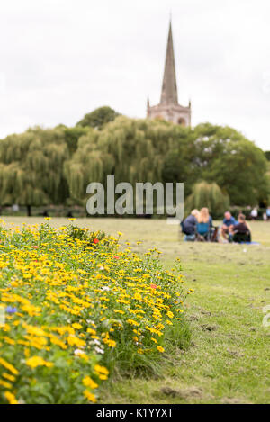 Vordergrund der Wiese Blumen und der Familie bei einem Picknick auf einer geschnittenen Gras Park mit einem Kirchturm und Willow Bäume in der Ferne. Stockfoto